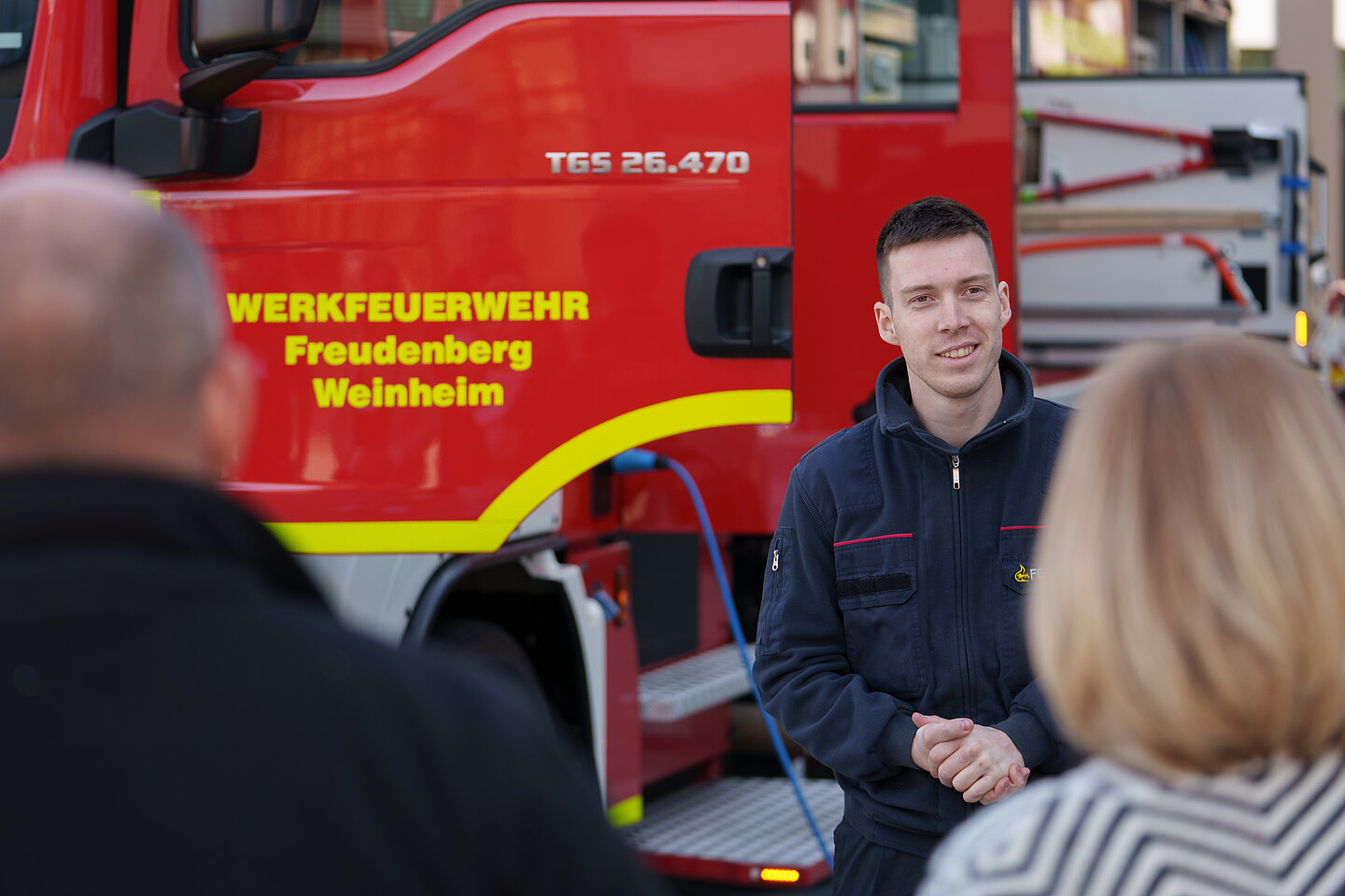 Ein Werkfeuerwehrmann in Uniform steht vor einem Werkfeuerwehrauto und unterhält sich mit zwei Menschen.