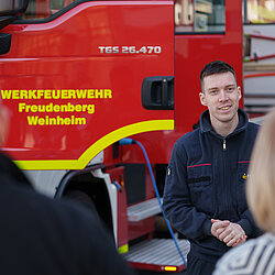 Ein Werkfeuerwehrmann in Uniform steht vor einem Werkfeuerwehrauto und unterhält sich mit zwei Menschen.
