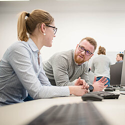 Zwei Auszubildende sitzen in einem Computerraum und schauen sich an.