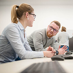 Zwei Auszubildende sitzen in einem Computerraum und schauen sich an.