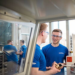 Zwei männliche Auszubildende in blauen T-Shirts und Arbeitsbrillen stehen gemeinsam vor einer Maschine, im Hintergrund sind weitere Auszubildende.
