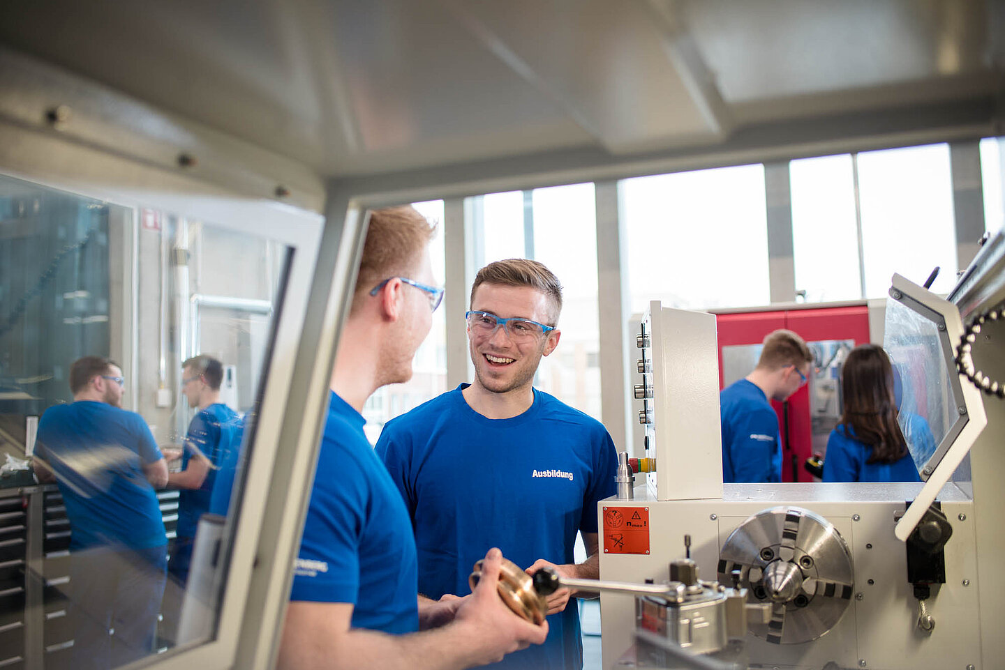Zwei männliche Auszubildende in blauen T-Shirts und Arbeitsbrillen stehen gemeinsam vor einer Maschine, im Hintergrund sind weitere Auszubildende.