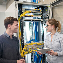 Zwei Auszubildende stehen sich gegenüber und sprechen miteinander vor einer technischen Umgebung.