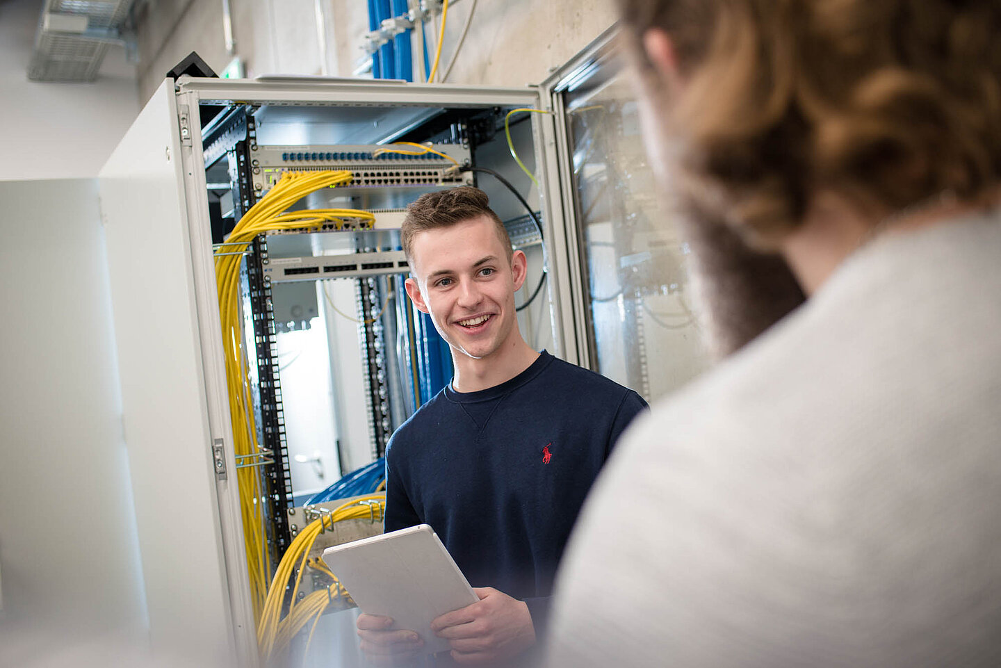 Ein Auszubildender steht vor technischer Umgebung, hält einen Block in der Hand und lächelt.